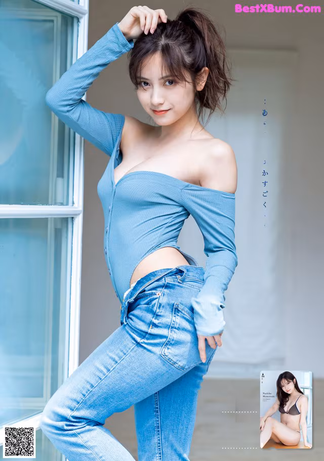 A woman in a blue top and jeans posing by a window.
