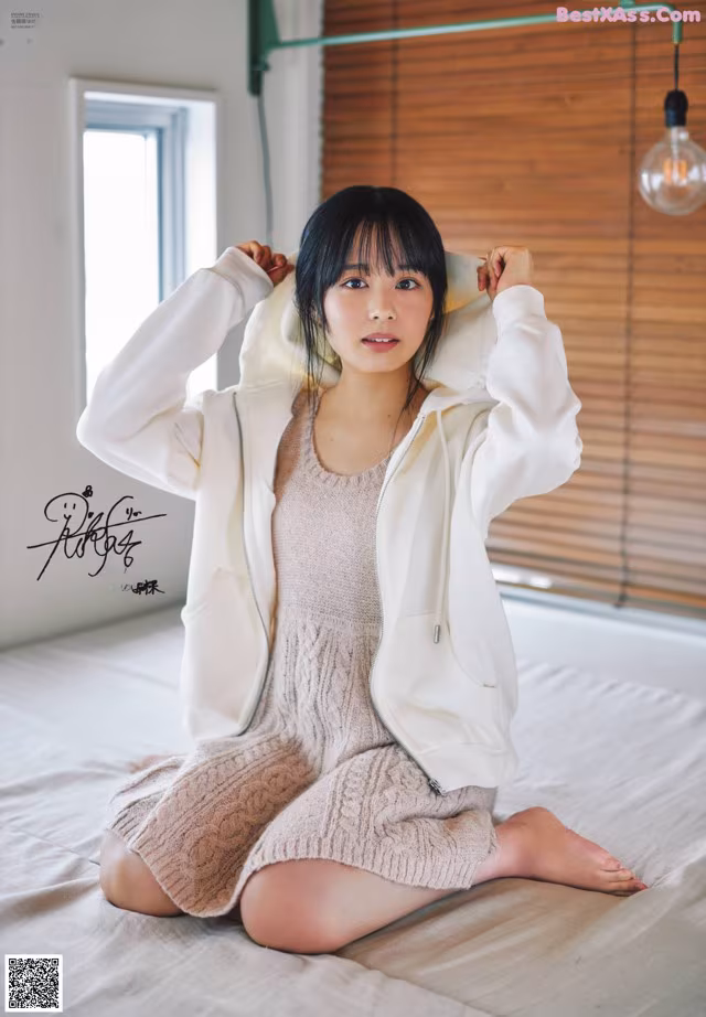 A woman sitting on a bed wearing a white jacket.